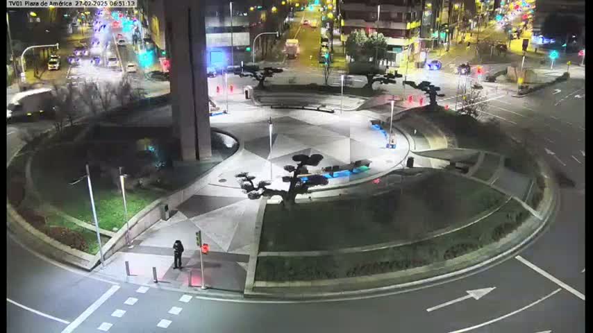 A nighttime aerial view of a circular plaza with landscaped areas and sparse traffic, illuminated by streetlights.