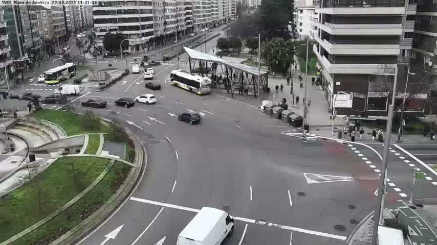 A city intersection with several cars and a bus, surrounded by buildings and landscaping, on a cloudy day.