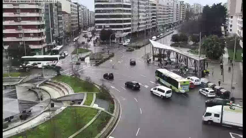 A wet, city intersection with several buses, cars, and pedestrians, surrounded by multi-story buildings.