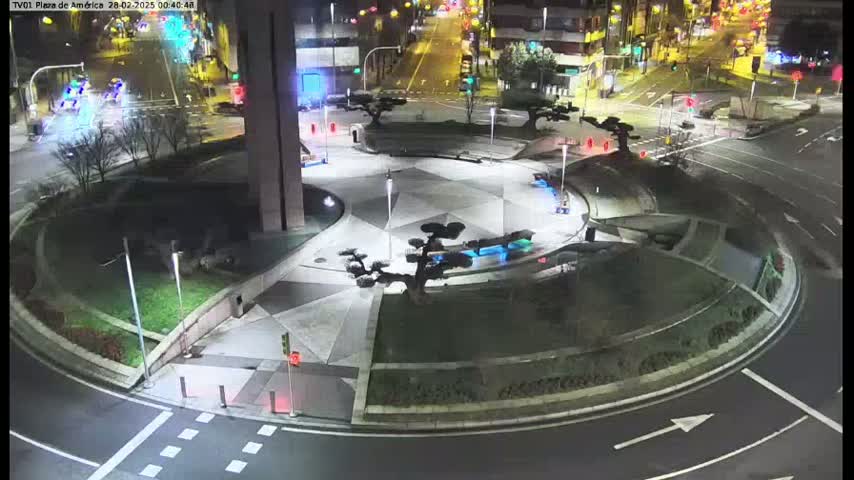 A nighttime aerial view of a circular plaza with landscaped areas and small trees, situated at a traffic intersection with cars visible on the roads.