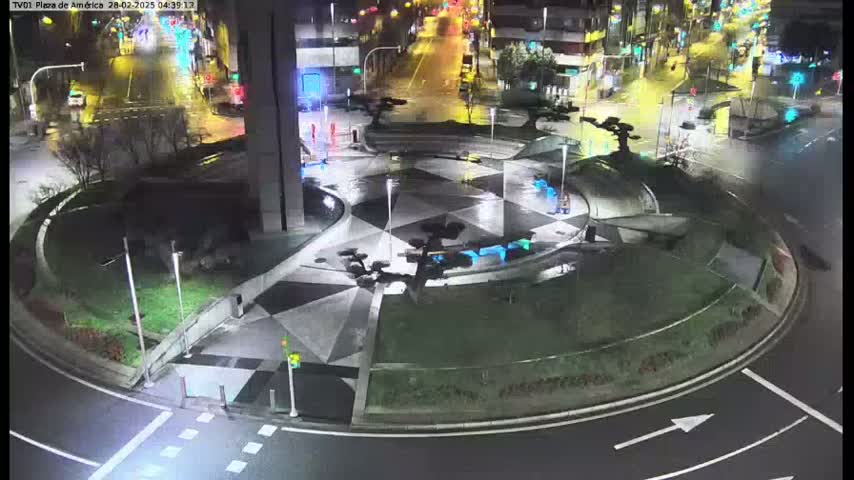 A nighttime aerial view shows a wet, geometrically patterned roundabout with sparse landscaping and minimal traffic.