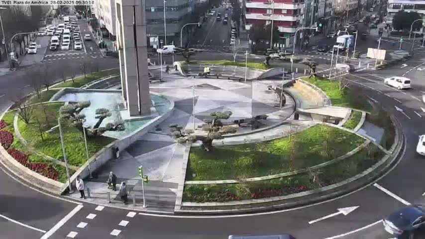 A landscaped traffic circle with a central plaza and fountain is surrounded by roadways with cars, under a mostly sunny sky.