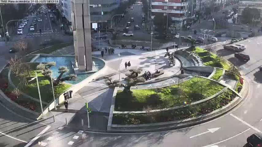 A sunny day reveals a circular plaza with landscaped gardens, water features, and a few pedestrians, surrounded by city streets with cars.
