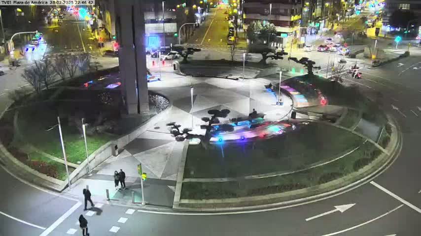 A nighttime aerial view of a circular plaza with landscaped areas and several pedestrians, surrounded by roadways with cars.