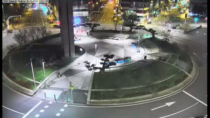 A nighttime aerial view shows a circular plaza with several small trees and geometric patterns in a paved area, surrounded by roads with minimal traffic.
