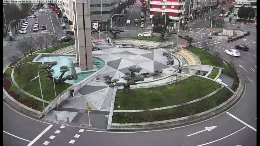 A paved roundabout with landscaped areas, including small trees and a water feature, is surrounded by city streets with several cars visible; the weather appears overcast.