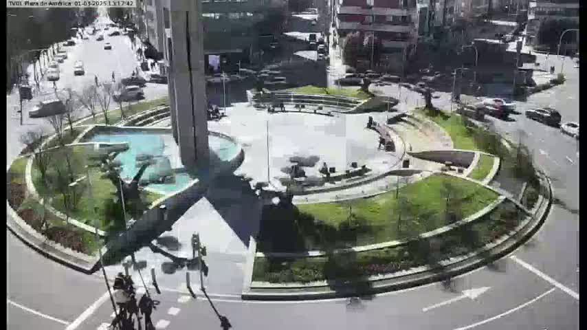 A circular plaza with landscaped areas, fountains, and seating surrounds a central monument, viewed from above on a sunny day with cars driving on surrounding streets.