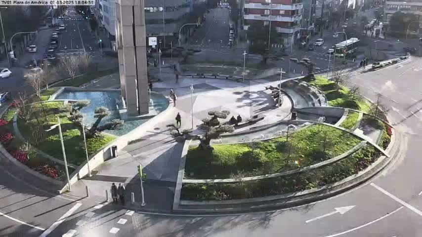 A sunny day reveals a circular plaza with a central fountain, landscaping, and a few pedestrians, situated within a city streetscape.