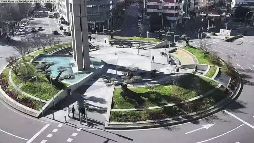 A sunny day shows a landscaped traffic circle with a fountain, several trees, and a few pedestrians.
