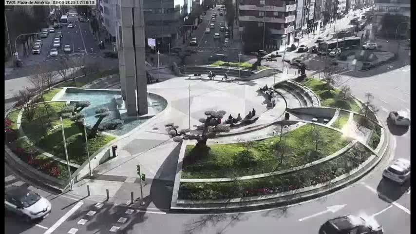 A sunny day reveals a circular plaza with a fountain, landscaping, and several people sitting in the center, surrounded by a roadway with cars.