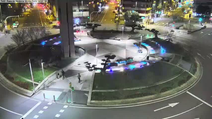 A nighttime aerial view of a circular plaza with landscaped areas, water features, and several pedestrians, surrounded by roadways and buildings.