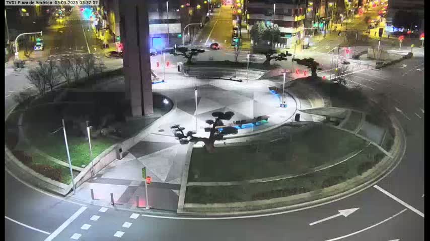 An illuminated roundabout at night, surrounded by landscaping and buildings, with a few cars visible in the distance.