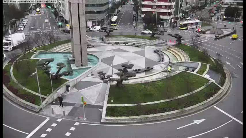 A roundabout in a city, featuring landscaped areas with small trees and a central fountain, surrounded by roads with light traffic under an overcast sky.
