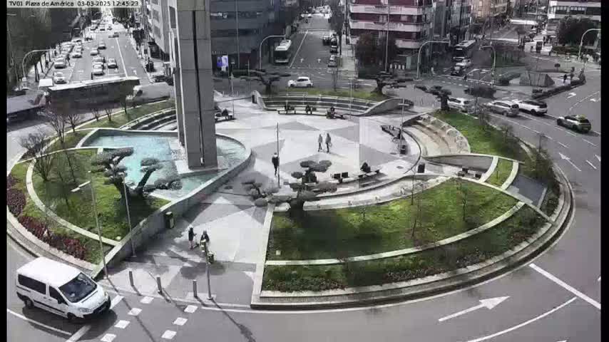 A landscaped roundabout with a central fountain and several people walking around, surrounded by city streets with cars driving on a sunny day.