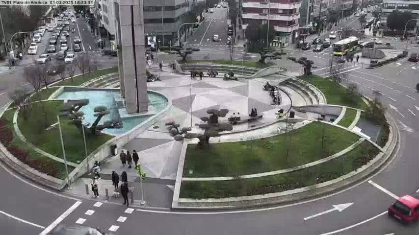 A circular plaza with a fountain and landscaping is situated in the center of a roundabout with several cars and pedestrians visible on a partly cloudy day.