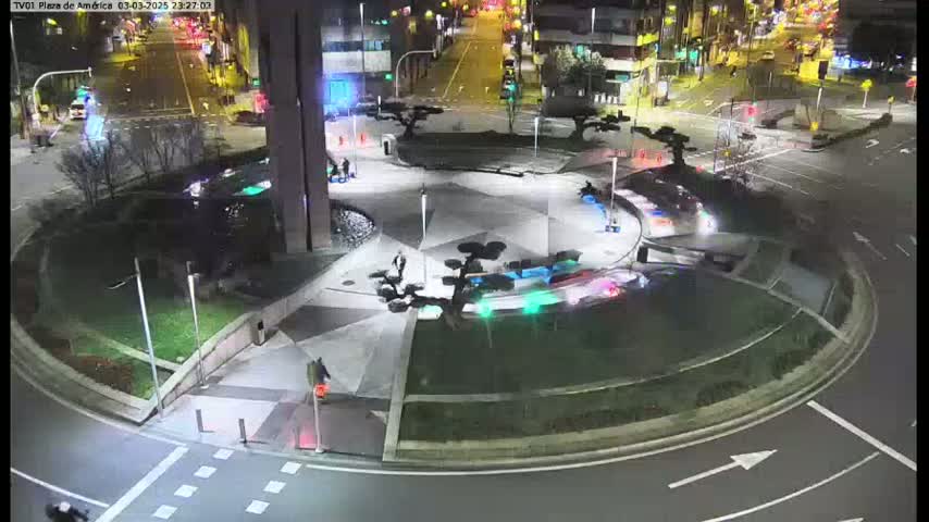 A nighttime view of a roundabout with minimal traffic, surrounded by landscaping and buildings, under clear skies.