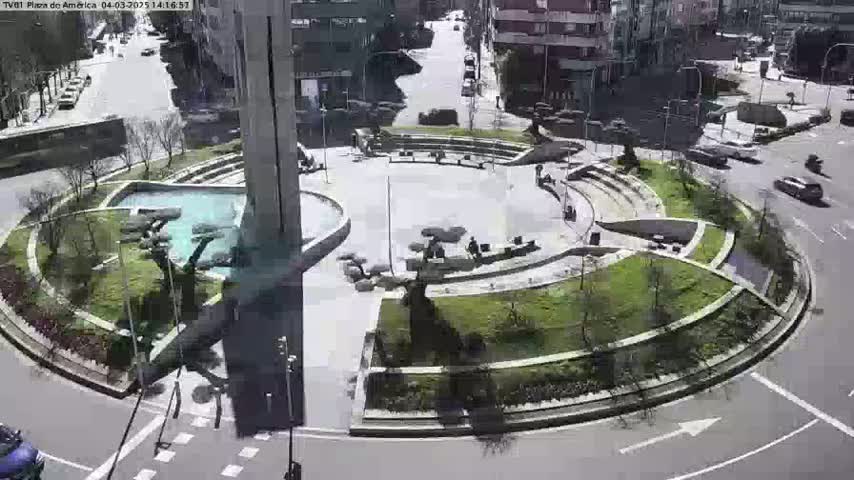 A sunny day reveals a circular plaza with landscaping, a central fountain, seating areas, and surrounding buildings, all viewed from above.