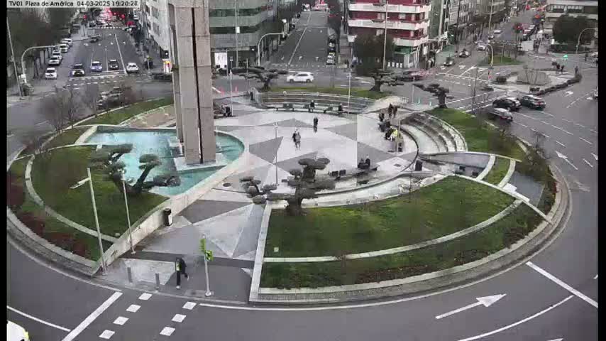 A circular plaza with a central fountain, landscaping, and pedestrian walkways is surrounded by a roadway with cars and buildings in the background, on a mostly cloudy day.