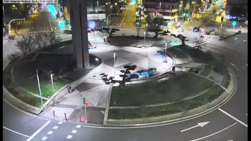 A nighttime aerial view shows a circular plaza with landscaped areas and trees, surrounded by roadways and traffic lights.