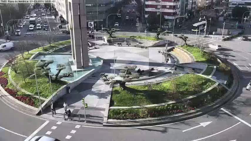 A circular plaza with landscaping, a water feature, and several small trees is situated in the center of a roundabout, with cars visible on the surrounding streets in bright, sunny conditions.