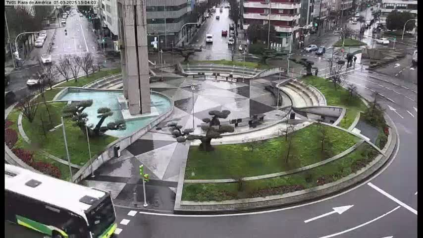 A wet, circular plaza with landscaping, a fountain, and a tall building in the background is viewed from above.