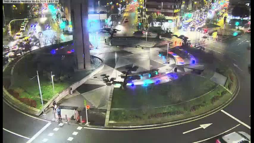 A nighttime aerial view of a circular plaza with a geometric pattern, surrounded by roadways with cars, and a few pedestrians near a crosswalk.