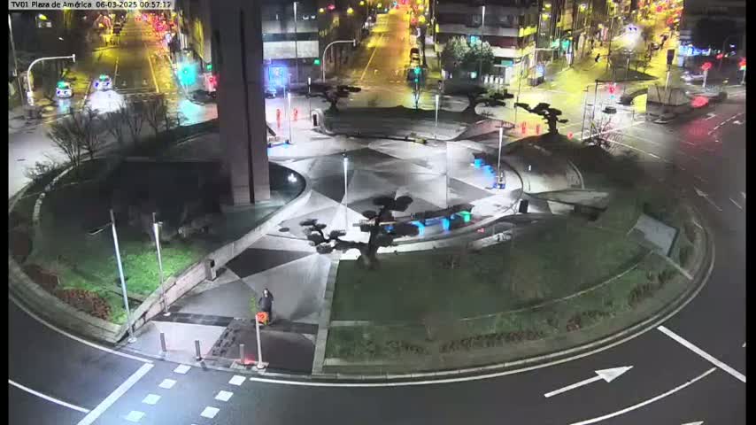 A nighttime aerial view of a circular plaza with geometric landscaping, illuminated by streetlights, and a few cars visible on surrounding roads.