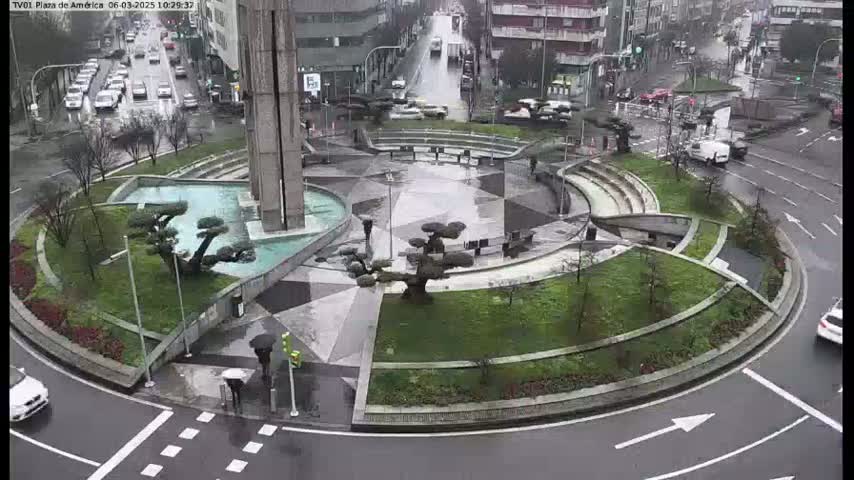 A rain-slicked roundabout features a central plaza with landscaping, a fountain, and several small trees, surrounded by a circular road with cars.