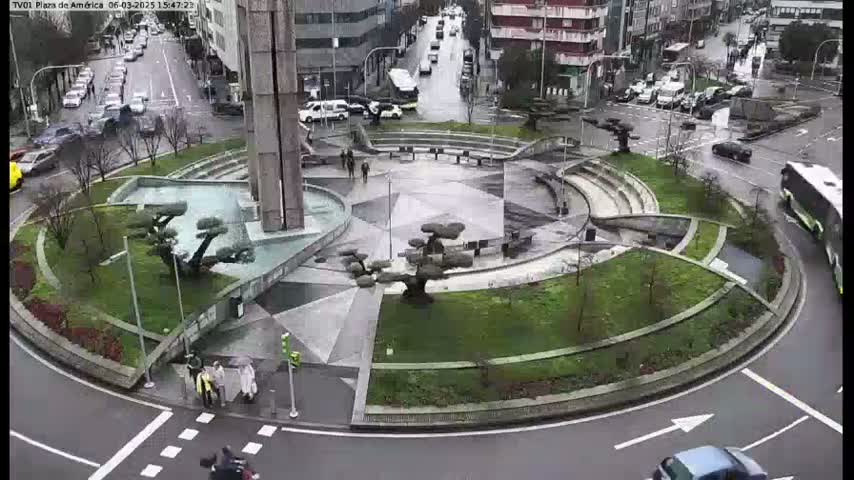A rain-soaked roundabout features a modern, geometric plaza with landscaping, a central fountain, and several pedestrians.