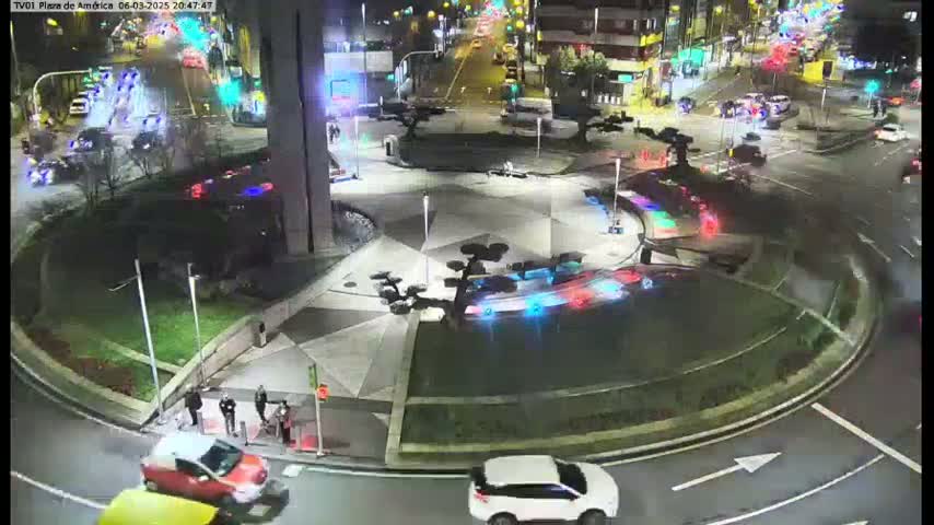 At night, several cars are navigating a roundabout in a city, with some pedestrians visible near a building.
