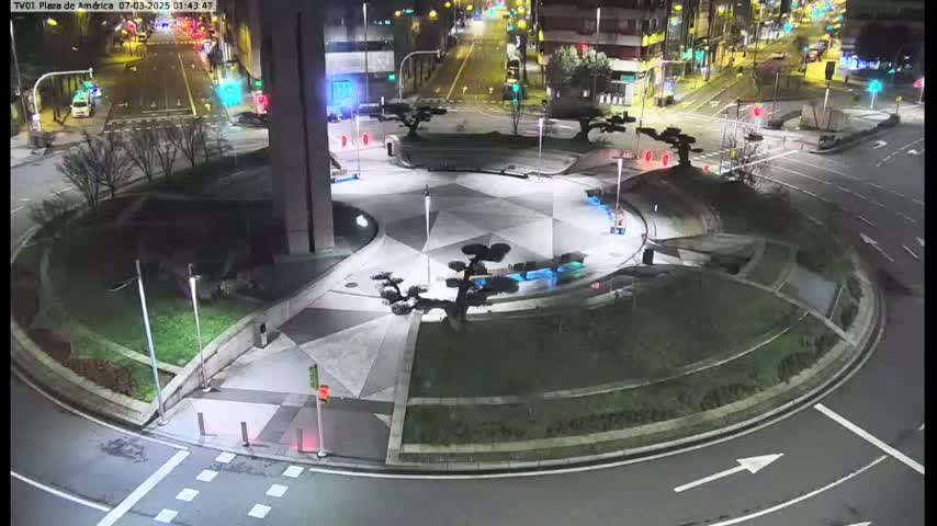 A nighttime aerial view shows a circular plaza with landscaping and a few cars on roads surrounding it.