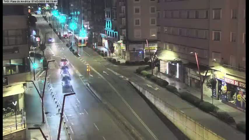 A nighttime, wet street scene shows several cars driving on a multi-lane road that passes buildings with streetlights illuminating the area.