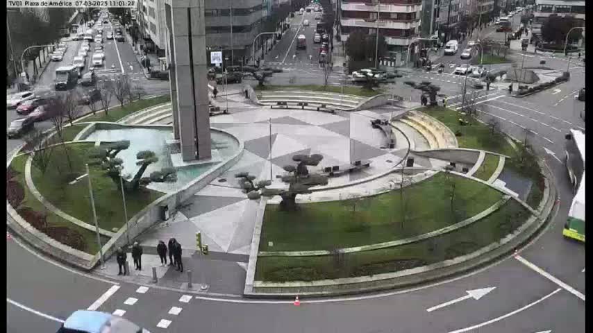 An aerial view of a circular plaza with a geometric stone design, water features, and landscaping, surrounded by city streets and buildings on an overcast day.
