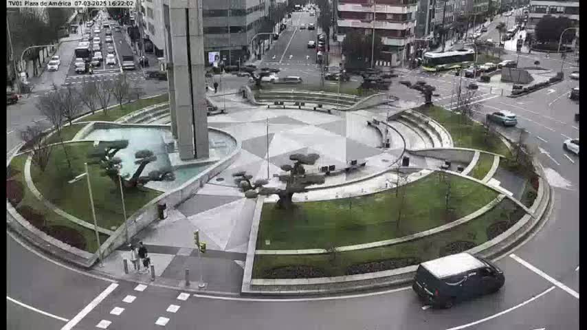 A circular plaza with landscaped gardens and a central water feature is surrounded by roads with cars, under an overcast sky.