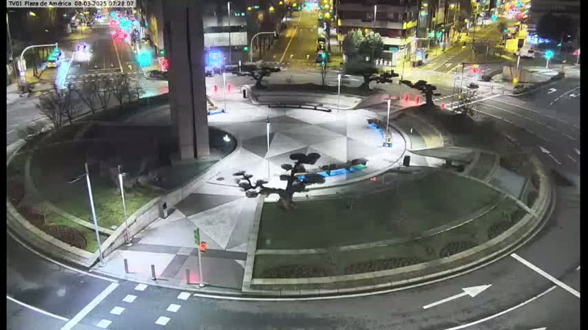 A nighttime aerial view of a circular plaza with landscaped areas and small trees, situated at a crossroads with cars visible on the surrounding streets.