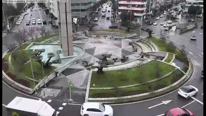 A roundabout with a central fountain and landscaping is surrounded by wet roads with several cars driving on a cloudy day.