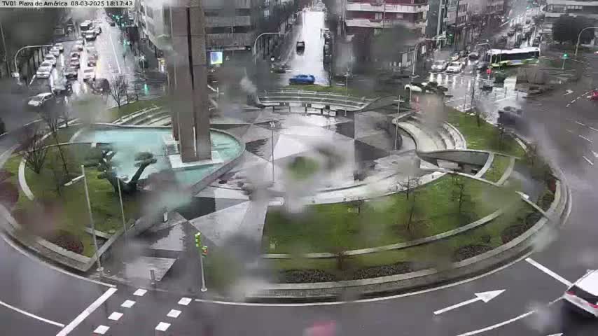 A rain-soaked roundabout with a central plaza and surrounding landscaping is shown, with cars driving on the surrounding streets.
