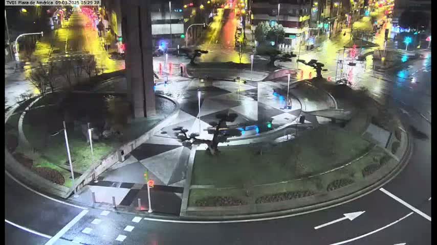 A wet, nighttime aerial view of a roundabout with geometric landscaping and small trees, surrounded by roadways with sparse traffic.