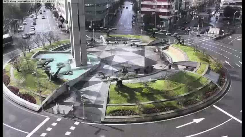 A landscaped roundabout with a geometric, patterned center is surrounded by roads with cars, under an overcast sky.