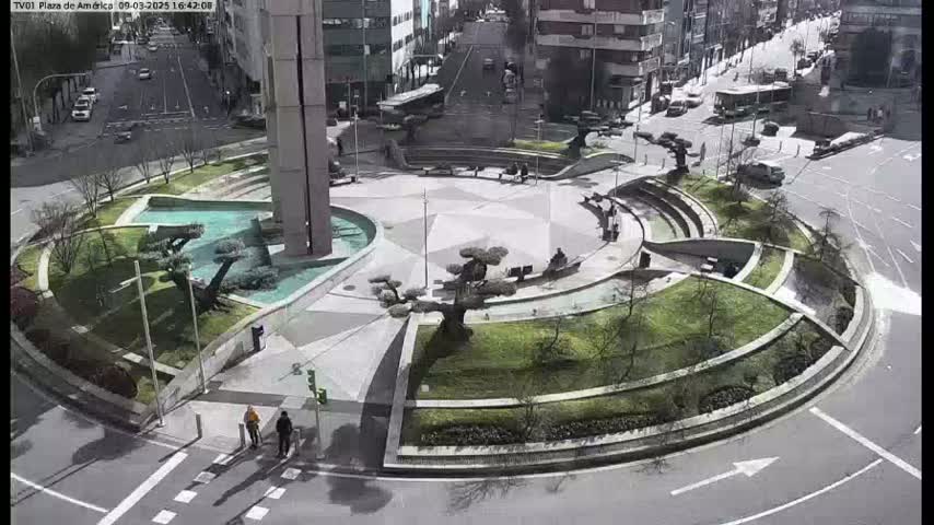 An aerial view shows a circular plaza with landscaped gardens, fountains, and a tall building in the background on a partly sunny day.