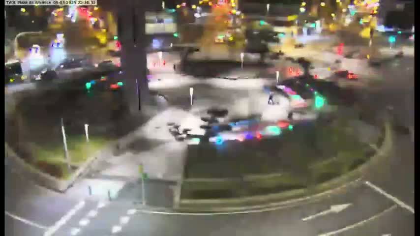 A night-time, high-angle view shows a roundabout with cars and pedestrians moving around it under artificial light.
