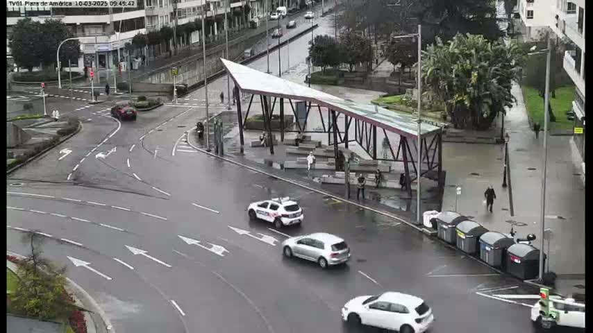 A wet city intersection with several cars, a modern bus shelter, and pedestrians, under an overcast sky.