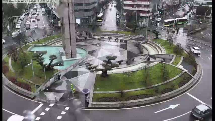 A wet, circular plaza with landscaping and a fountain is surrounded by a roadway with cars driving in the rain.