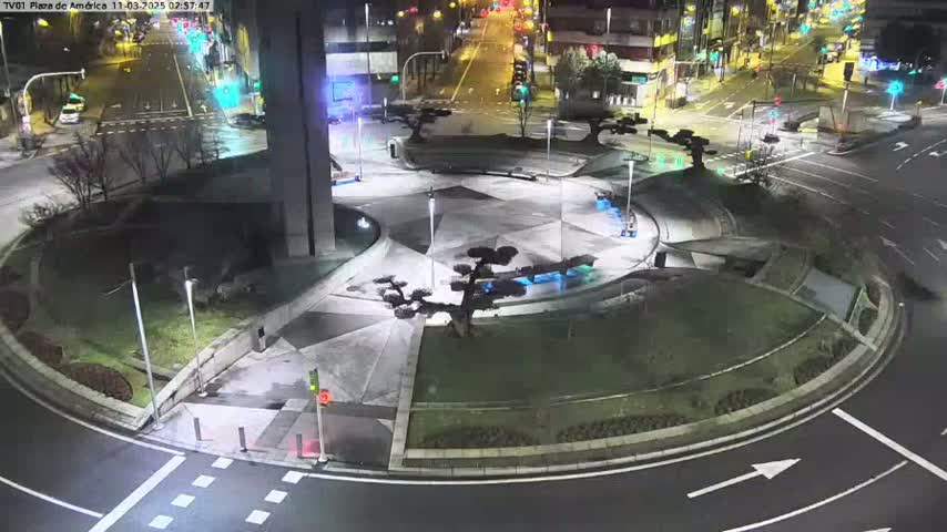 A nighttime aerial view of a landscaped roundabout with a central, geometric plaza featuring several small trees, surrounded by roadways with cars and traffic signals under slightly wet conditions.