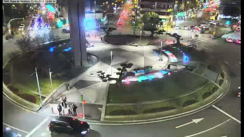 A nighttime aerial view of a roundabout with cars and pedestrians, illuminated by streetlights, in a plaza setting with landscaped areas.