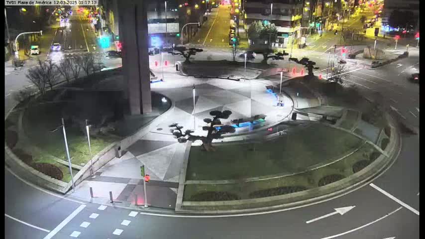 A nighttime view of a circular plaza with several small trees, surrounded by roadways with sparse traffic.