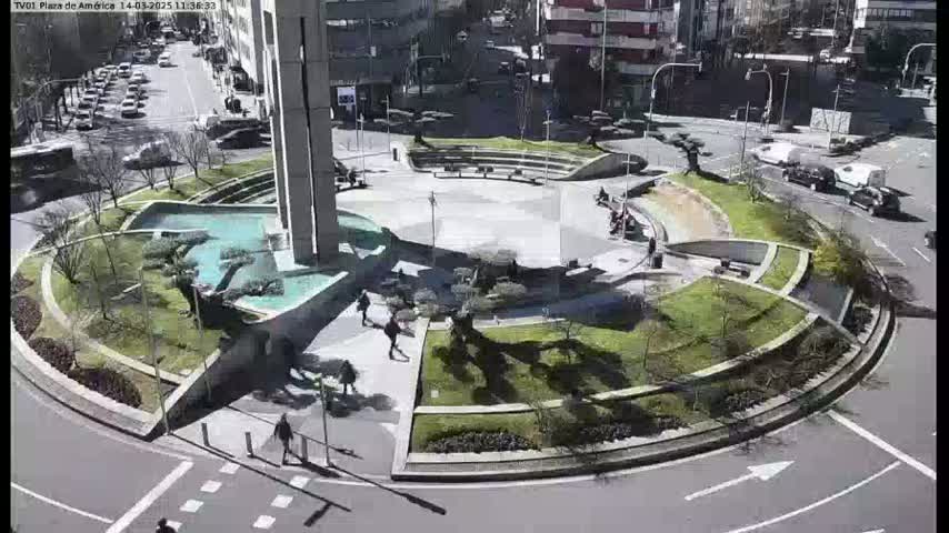 A sunny day reveals a circular plaza with landscaping, a central fountain, and several pedestrians, surrounded by a roadway with cars.