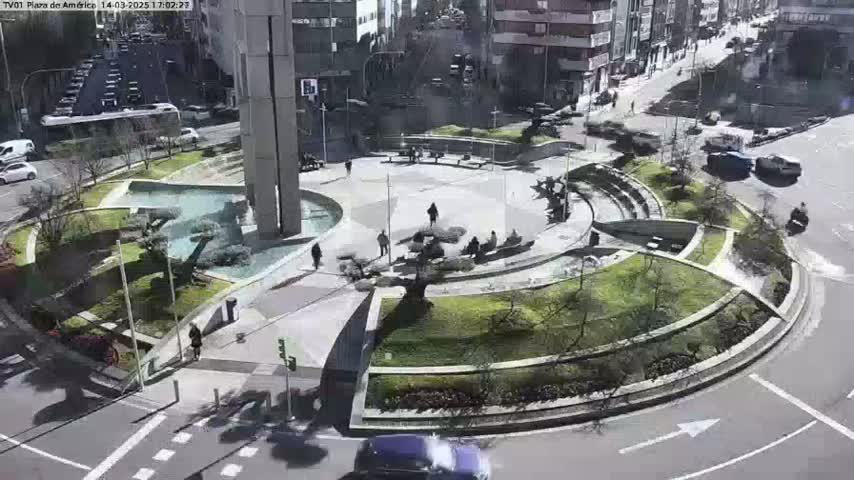 A circular plaza with landscaping, a fountain, and several people is situated in the center of a roadway on a sunny day.