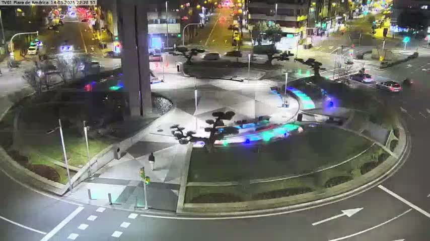 A nighttime aerial view of a roundabout with several cars and illuminated landscaping features.