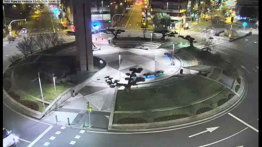 A nighttime aerial view shows a circular plaza with geometric landscaping and trees, situated at a traffic intersection with cars visible on the roads.